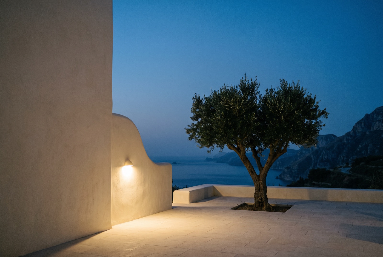 A man seen from behind, cup of coffee in hand, on the wrought-iron balcony of a white house in Ojén at dawn, looking down the valley towards the sea as the first light touches the village rooftops.