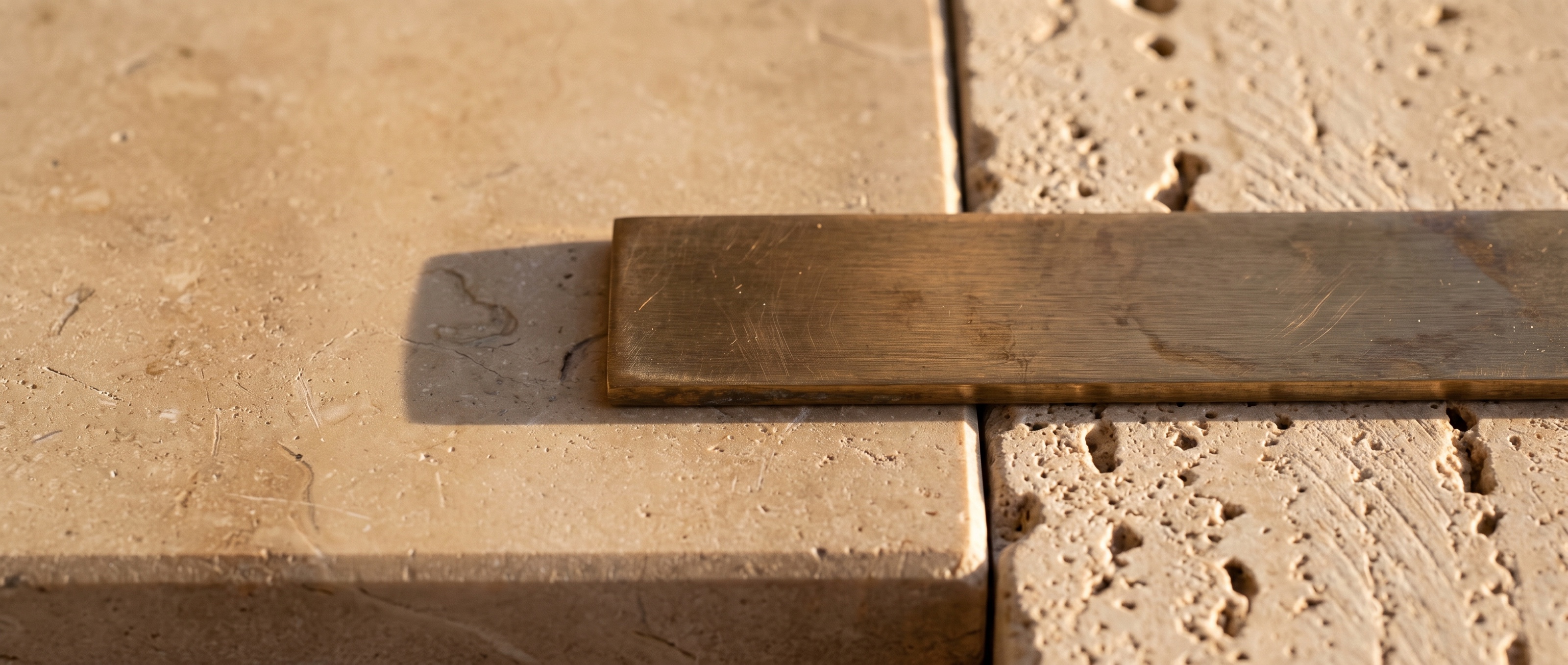 Primerísimo plano de piedra caliza, travertino y bronce cepillado bajo luz mediterránea cálida, con sombras suaves.