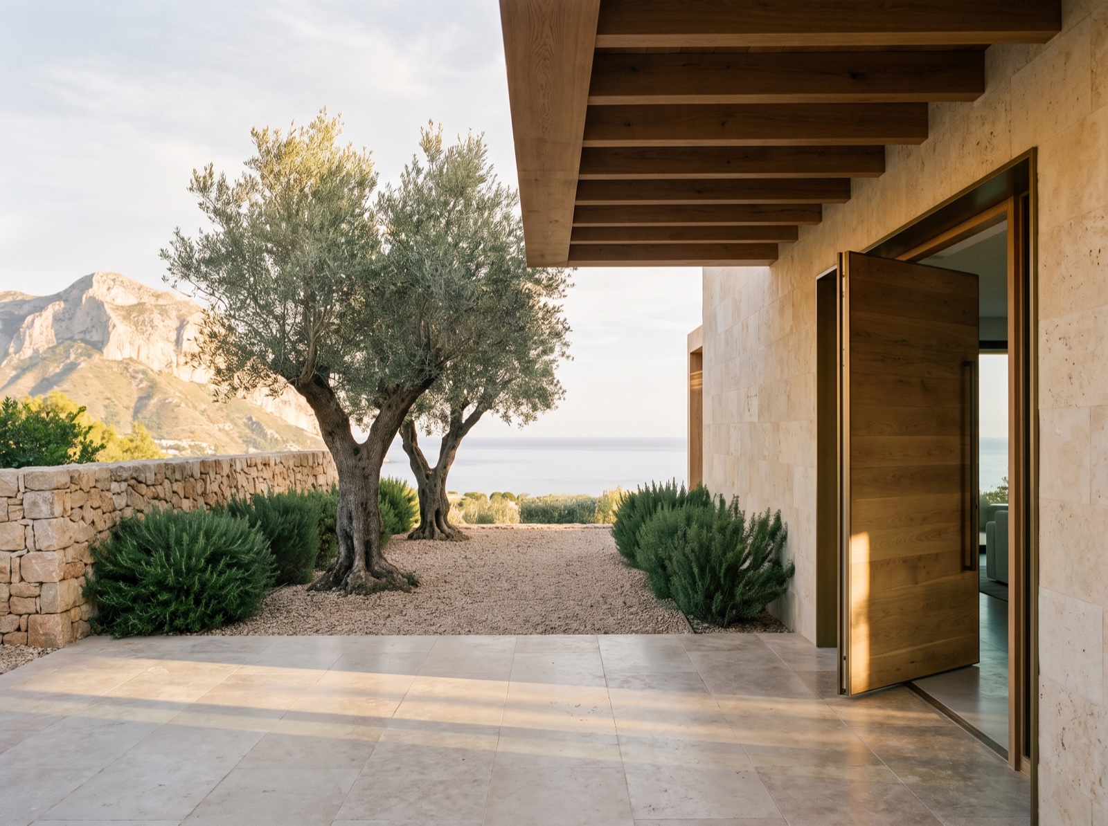 Entrance courtyard of a contemporary Marbella villa: dry-stone wall, olive trees, an oak door left ajar and the Mediterranean beyond, with the mountain to the left
